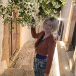 Ila walks down a quiet, illuminated side alley in Lindos wearing an elegant yet comfortable outfit (flat sandals and a light jacket mix), proving the ideal dress code for the uneven cobblestone streets.