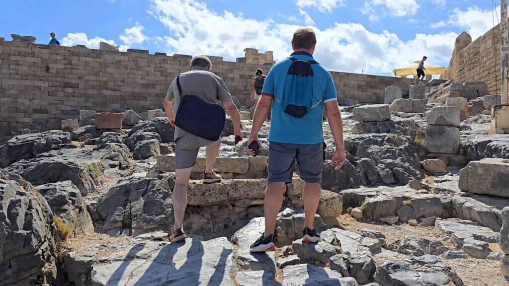 Acropolis of Lindos Guide: First-Hand Tips, Best Time to Visit, and History 5 Gabriel and Pietro ascending the steep, irregular stone steps of the Acropolis of Lindos, demonstrating the climb is manageable with suitable footwear, framed by the medieval Knights' Fortress wall.