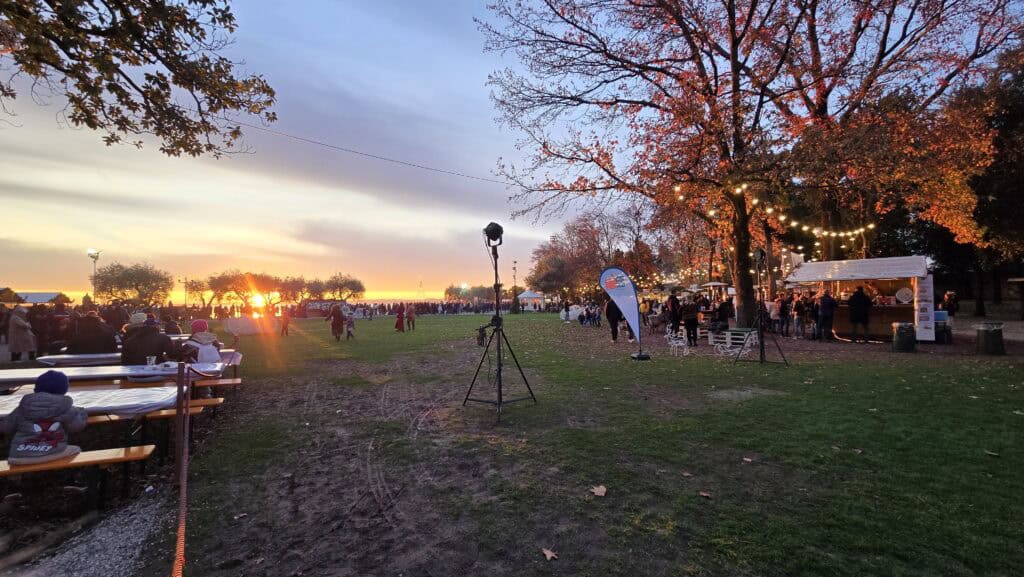 A wide shot of the Bardolino Christmas Market at sunset on Lake Garda. The scene features people sitting at picnic tables in a grassy park, festive string lights illuminating the food stalls, and the sun setting brightly over the lake in the background. The atmosphere is lively and festive, confirming the town's enchanting holiday experience.