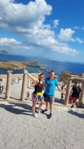 Ila and Gabriel standing at the Acropolis of Lindos at 11:30 AM on October 4th, enjoying the comfortable weather for sightseeing, without the peak-summer heat.