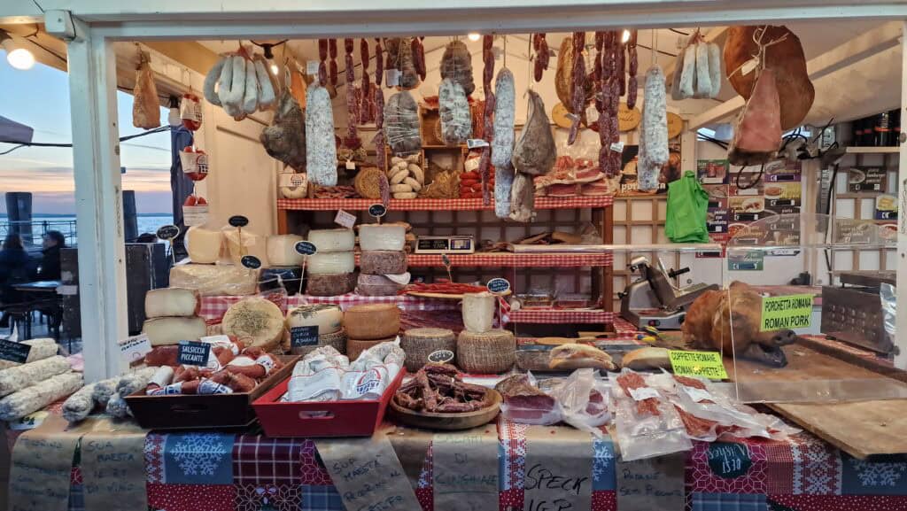 A market stall selling traditional Italian cured meats like salami and prosciutto at the Lazise Christmas Market on Lake Garda in winter.