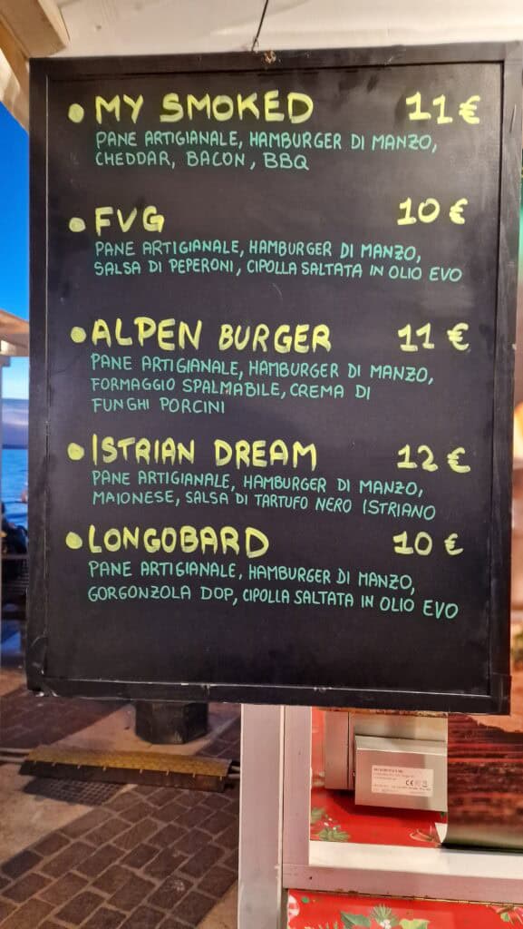 A stall selling large, hot sandwiches (panini) with visible prices at the Lazise Christmas Market, enjoyed on improvised lakeside benches at sunset.