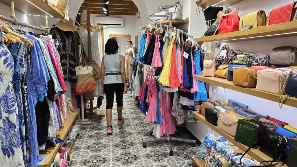 Interior view of a Lindos boutique with racks of colorful summer clothing, dresses, woven bags, and shelves displaying stylish handbags and accessories.