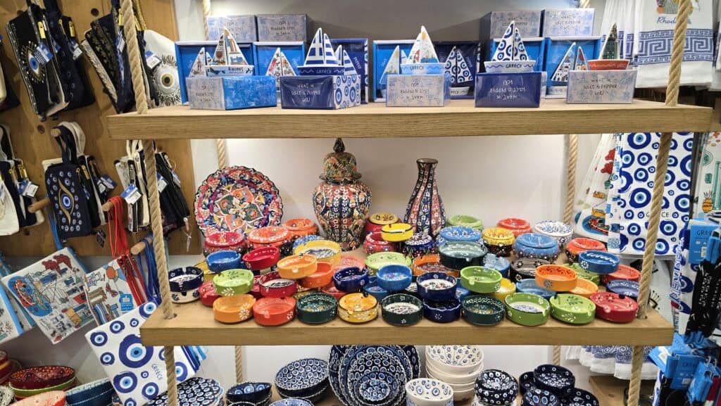 Display of colorful Greek ceramic bowls and traditional souvenir items in a Lindos shop, featuring blues, greens, reds, and oranges on wooden shelves.