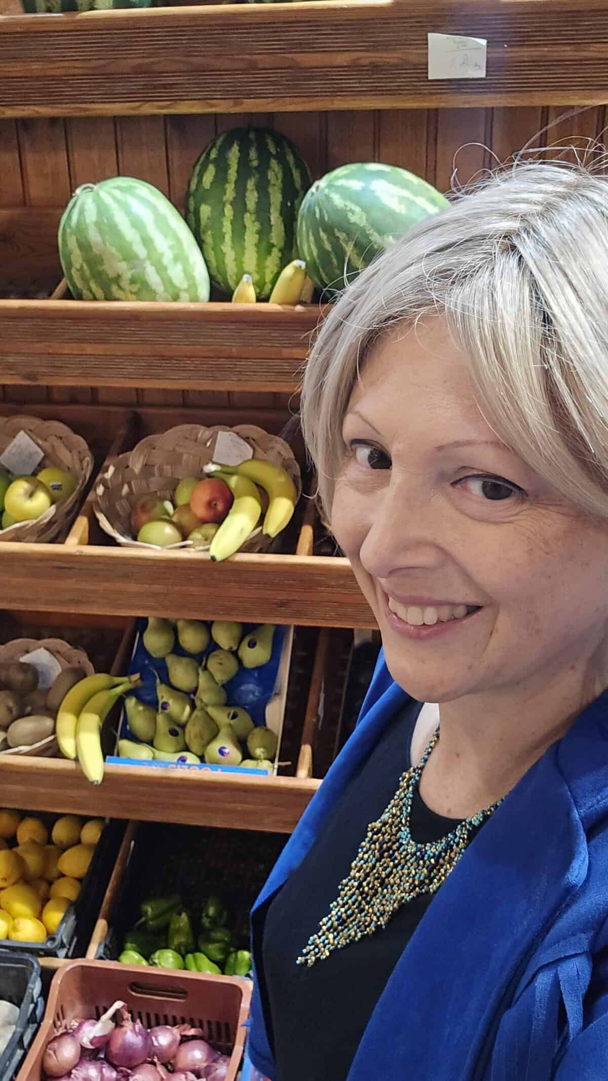 Ila smiles in front of a colorful fresh fruit stall (watermelons, bananas, lemons) in the main shopping street of Lindos, Rhodes, highlighting the village's vibrant atmosphere.