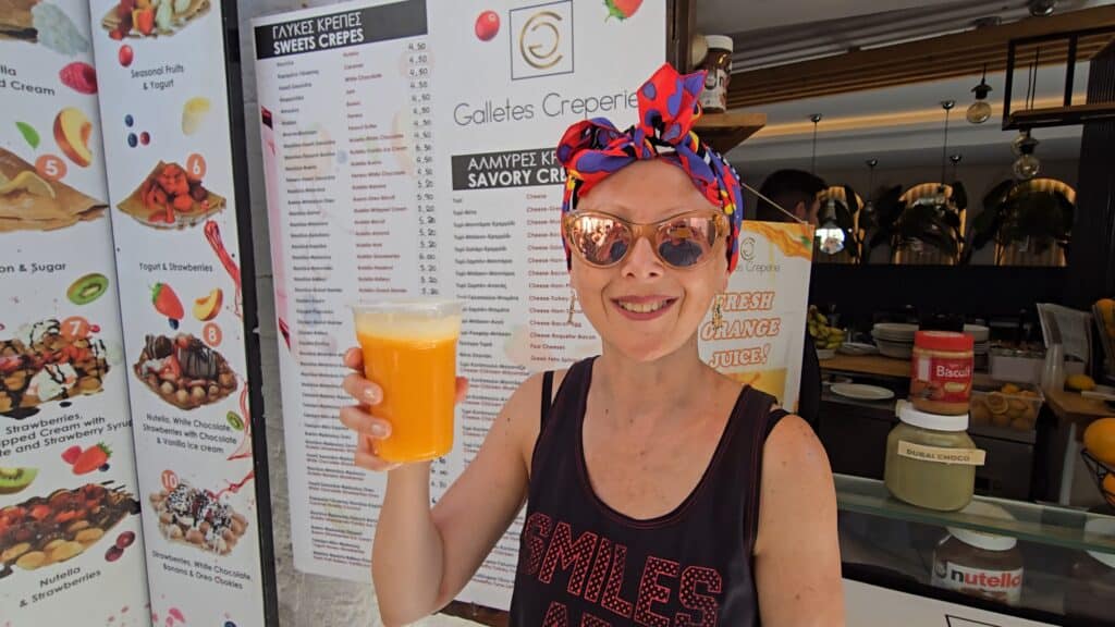 Lindos, Rhodes: The Complete Guide to What to See, Acropolis, and the White Village's Beaches 6 Ila enjoying a fresh orange juice at a Lindos street food kiosk, with the menu board for crepes (around €5) visible in the background, offering a low-cost, quick refreshment option after visiting the Acropolis.
