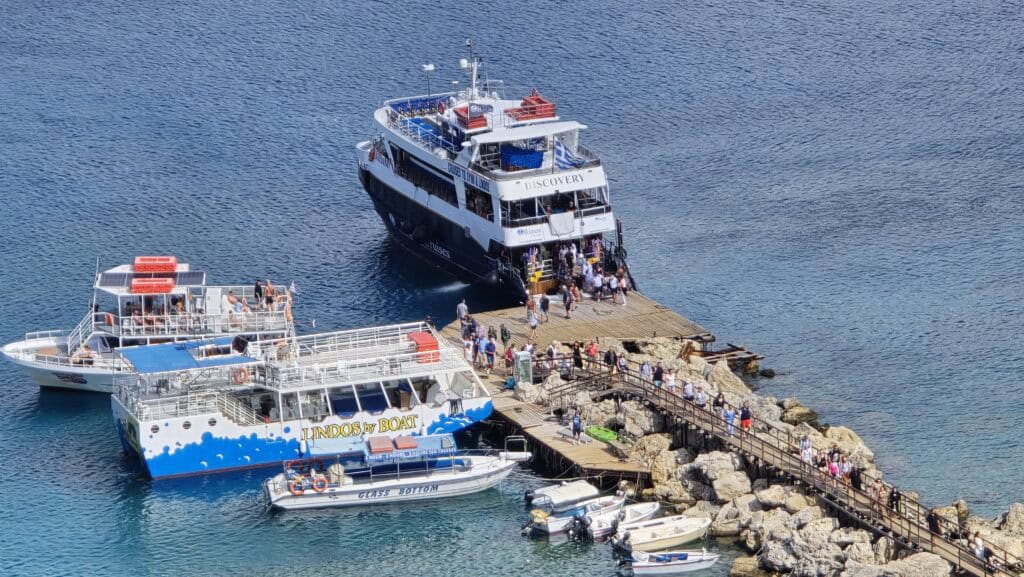 Lindos, Rhodes: The Complete Guide to What to See, Acropolis, and the White Village's Beaches 8 Tourist boats, including a glass bottom boat and large excursion ferries, docked at the stone jetty near Lindos Main Beach, ready for coastal tours. People boarding cruise ships at a busy harbor dock on a clear day.