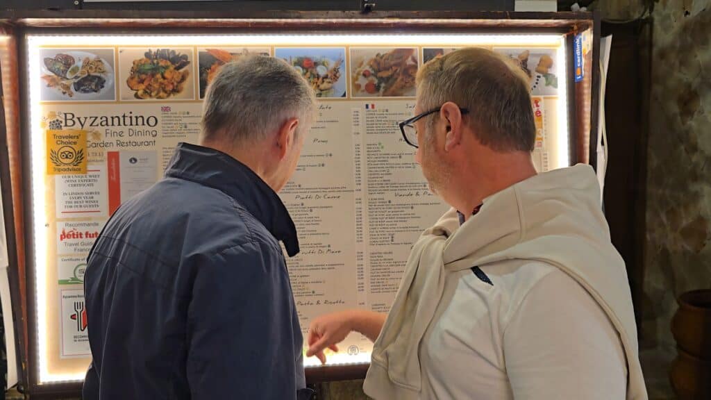 Two people (Gabriel and a companion) examine a brightly lit outdoor menu board for the Byzantino Fine Dining Garden Restaurant in Lindos, discussing typical dishes and prices.