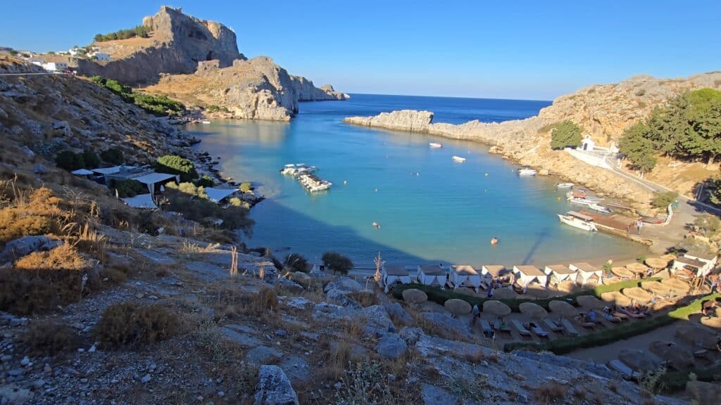 St. Paul's Bay in Lindos: a stunning, heart-shaped cove with turquoise water, overlooked by the majestic Acropolis atop the hill and a small white church on the right.
