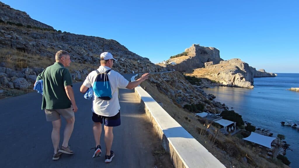 Gabriel and his travel companion walking on the stone pathway leading uphill from St. Paul's Bay towards the white historic center of Lindos, demonstrating the quick, scenic walk to the village.