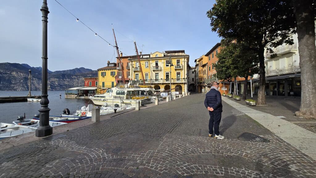 Gabriel stands alone, looking out over the completely deserted harbor of Malcesine, Lake Garda, on a sunny Sunday, December 7, 2025. The colorful historic buildings and moored boats contrast with the absence of people, illustrating the extreme quietude of the town in winter.