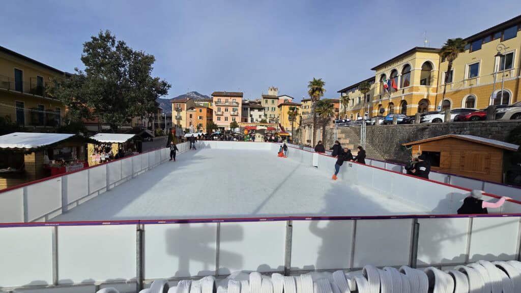 A panoramic view of the lively outdoor ice skating rink set up in the internal piazza of Malcesine, Lake Garda. People are skating and market stalls are visible, surrounded by colorful historic buildings. The location, away from the lakefront, emphasizes the quiet nature of Malcesine during the low season.