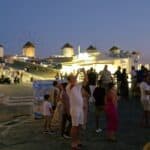 The famous Mykonos Windmills (Kato Mili) at sunset, showing the popular free viewpoint crowded with people, yet still offering space to move and enjoy the view.