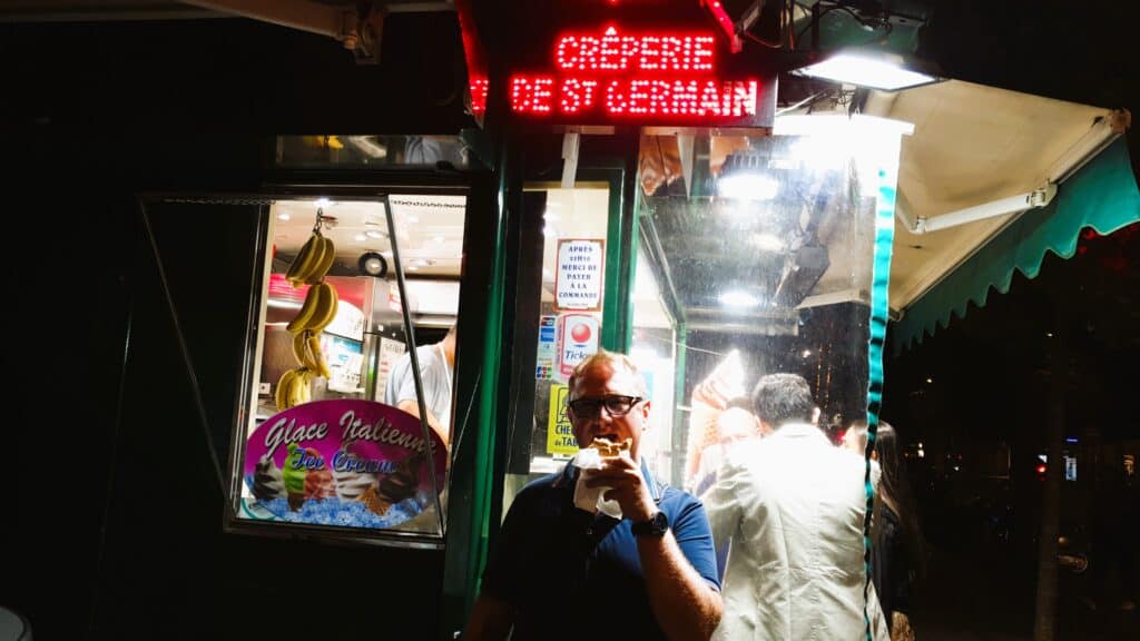 Parisian street food kiosk "Crêpes de Saint-Germain" illuminated in red with Gabriel enjoying the authentic experience.