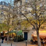 Parisian street with a classic café offering covered outdoor seating in November, highlighting bare trees and typical architecture.