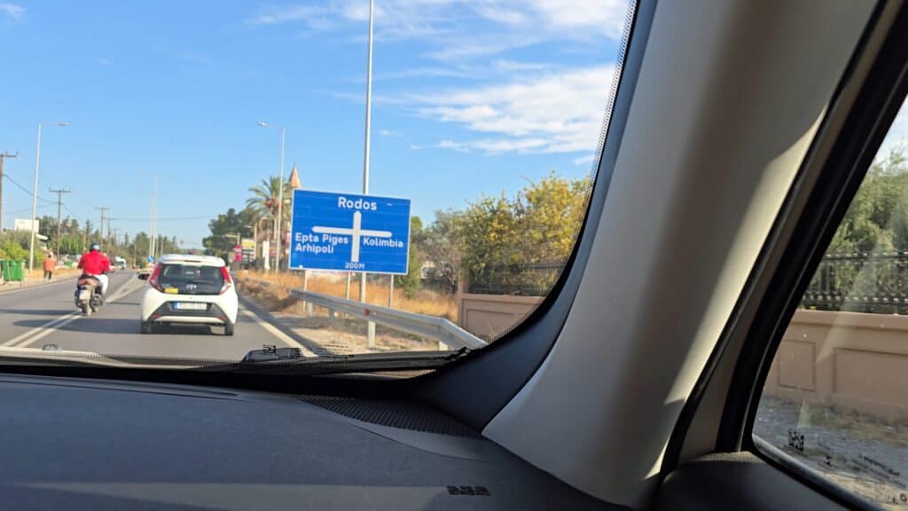 View from Gabriel and Ila's rented car on a wide, paved main road in Rhodes, showing good asphalt and traffic.