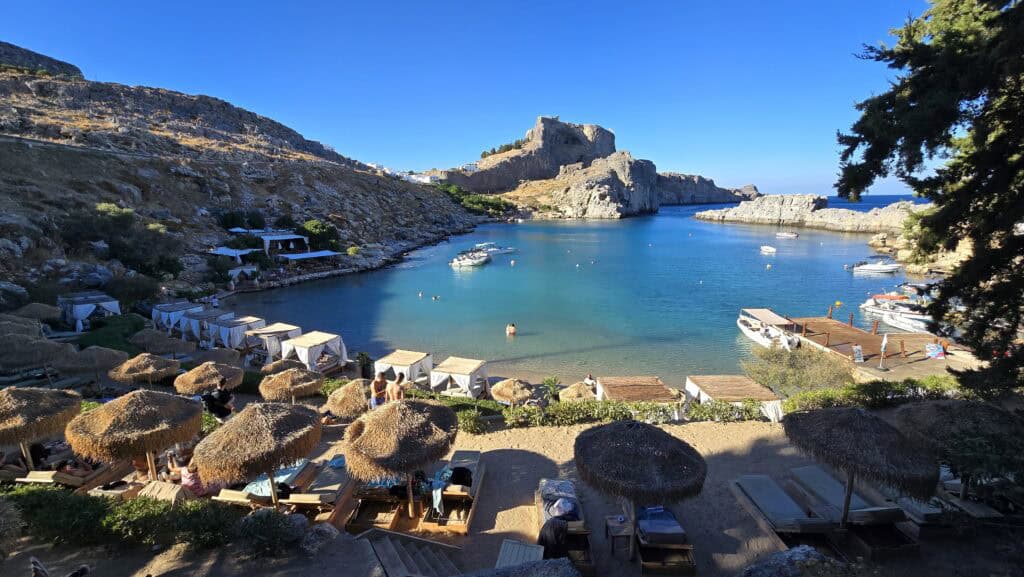 The view from the premium sunbeds at St. Paul's Bay: a true paradise scene showcasing the crystalline turquoise waters and the magnificent Acropolis of Lindos towering directly above the cove.