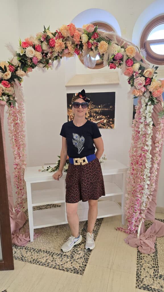 Majesty Reborn: Kallithea Springs Rhodes โ Italian Thermal Baths, Beaches & Architecture 6 Ila posing under a decorative wedding arch made of pink and white flowers and greenery, inside one of the white-walled rooms at Kallithea Springs.