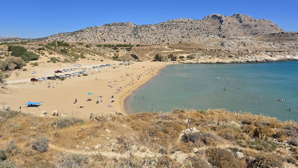 Panoramic view of Agathi Beach Rhodes, showing the golden sand, the large free parking area on the left, and the crystal-clear turquoise water in the sheltered bay.