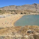 Panoramic view of Agathi Beach Rhodes, showing the golden sand, the large free parking area on the left, and the crystal-clear turquoise water in the sheltered bay.