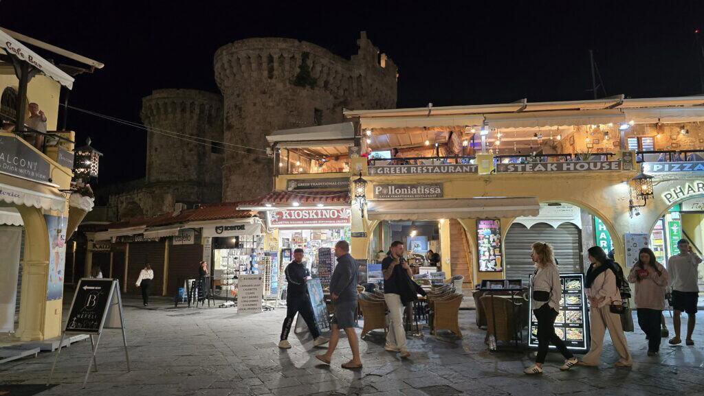 A lively view of Hippocrates Square (Ippokratous 26) at night in Rhodes Old Town, showing illuminated restaurants, including Polloniatissa Fish Taverna, and surrounding medieval walls.
