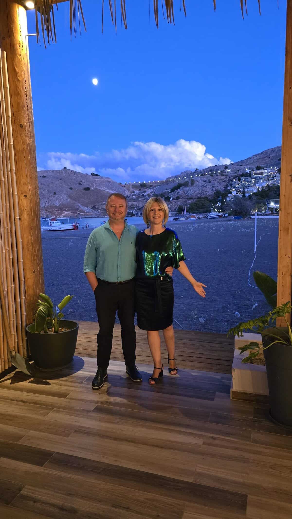What to Pack for Rhodes in September and October: Your Complete Guide 7 Ila and Gabriel dressed in elegant-sporty attire for a beach dinner at Vlicha Beach, Rhodes, with the bay and coastline in the background.