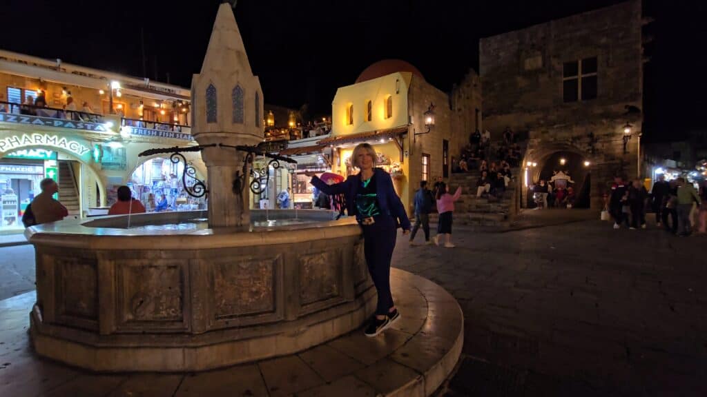 Ila standing in the illuminated Hippocrates Square (Platía Ippokrátous) in Rhodes Old Town at night, with the central fountain visible.
