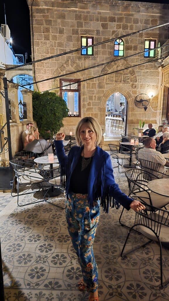 What to Pack for Rhodes in September and October: Your Complete Guide 5 Ila posing in a stylish blue jacket with fringe, floral trousers, and comfortable flat sandals at a charming outdoor bar in Lindos, Rhodes.