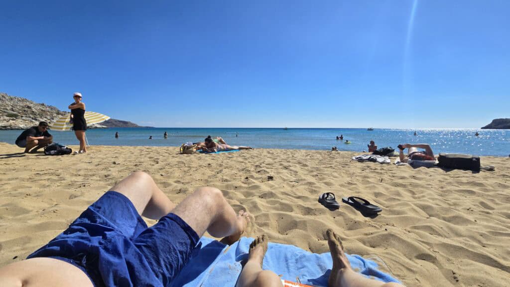 Ila and Gabriel relaxing on the soft golden sand of Agathi Beach Rhodes, with a first-person view of their legs and feet towards the clear Aegean Sea.