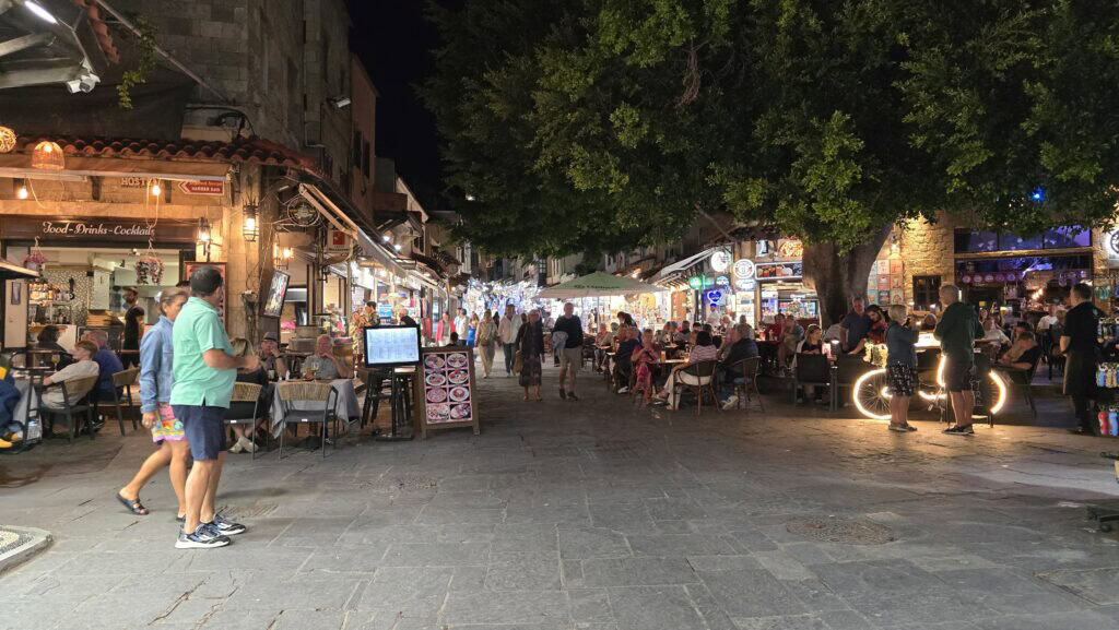 Lively street junction in Rhodes Old Town at night, showing people seated at outdoor cafe tables and strolling along the cobblestones.