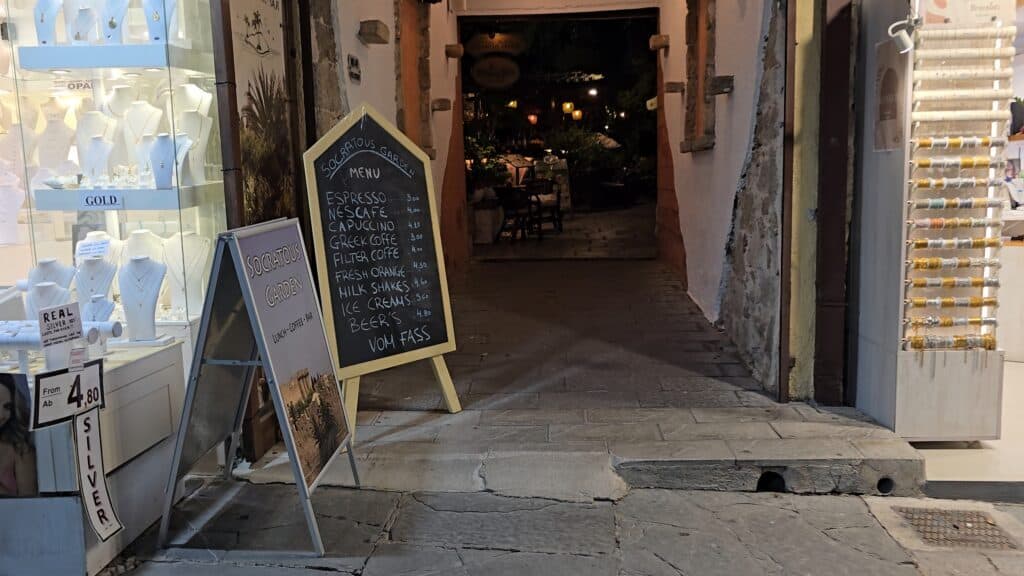 Hidden courtyard of Socratous Garden in Rhodes Old Town at night, showing soft lighting, tranquil seating, and a blackboard menu with prices.