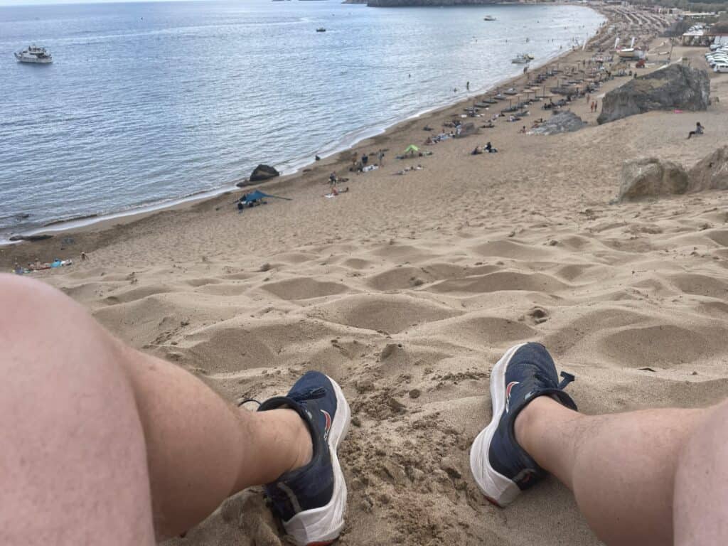 Tsambika Beach Rhodes: The Greek Flag Rock, Golden Sand & Crystal Clear Sea 3 A person (Gabriel) is sitting on the ridge of the steep sandy slope/dune at Tsambika Beach, with only their feet visible in the foreground, looking out over the panoramic view of the golden beach and crystal-clear sea below.