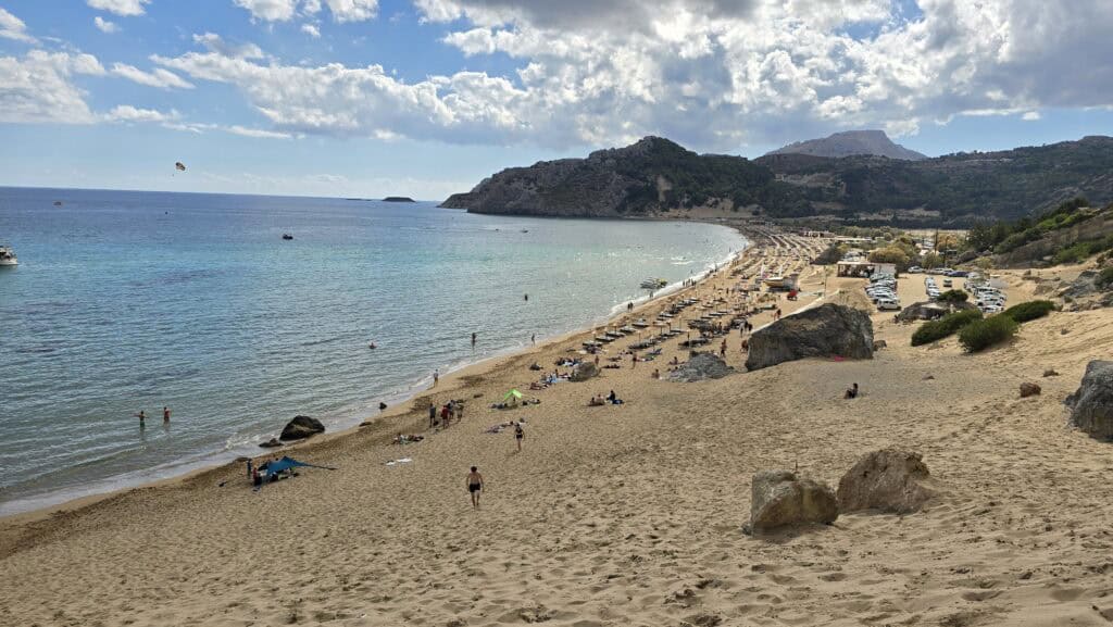 Tsambika Beach Rhodes: The Greek Flag Rock, Golden Sand & Crystal Clear Sea 1 Wide panoramic photo of Tsambika Beach taken from a high sandy vantage point, showing the full length of the golden sand, sunbeds, parking lot, clear turquoise water, and a partly cloudy sky with sunshine breaking through.