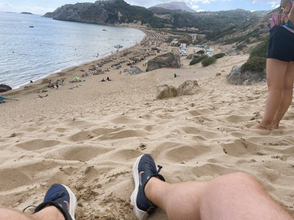 Tsambika Beach Rhodes: The Greek Flag Rock, Golden Sand & Crystal Clear Sea 4 High-angle photo taken from the top of the steep sandy slope at Tsambika Beach, showing the feet of two people (Ila and Gabriel) resting on the sand, overlooking the entire long beach and the turquoise sea below.