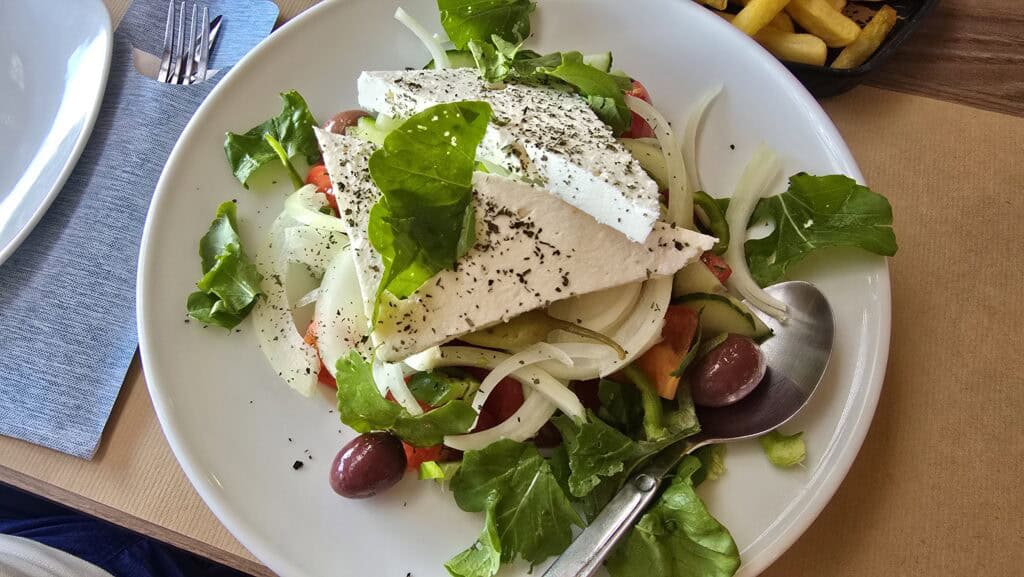 Tsambika Beach Rhodes: The Greek Flag Rock, Golden Sand & Crystal Clear Sea 6 Close-up photograph of a fresh Greek Salad (Horiatiki) featuring large slices of feta cheese seasoned with oregano, black olives, onions, cucumber, and tomatoes, served as part of a meal combo.