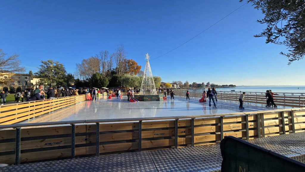 A sunny winter photo of the Bardolino outdoor ice skating rink, situated directly on the grassy park next to Lake Garda. The festive market stalls are visible. The cost of entry is typically €10.00.