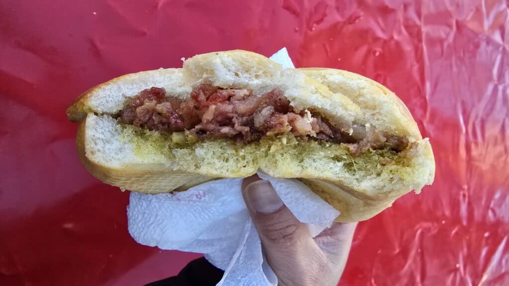 A close-up shot of a gourmet hot sandwich from the Bardolino Christmas Market, featuring sliced Cotechino sausage, hearty broccoli pesto, and a soft bun. The sandwich is held up near a market stall, ready to be eaten.
