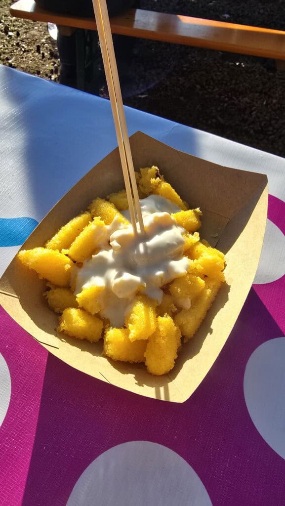 A close-up photograph of golden, crispy squares of fried polenta generously topped with creamy, melted Gorgonzola fondue, served in a paper cone from a Bardolino Christmas Market food stall.