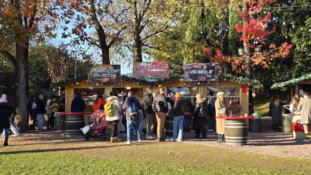 A food stall at the Bardolino Christmas Market, featuring clear signs for "Panino con la Salsiccia" and "Vin Brulée." Behind the stalls, the warm colors of the surrounding autumn trees confirm the late-fall, early-winter setting on Lake Garda.