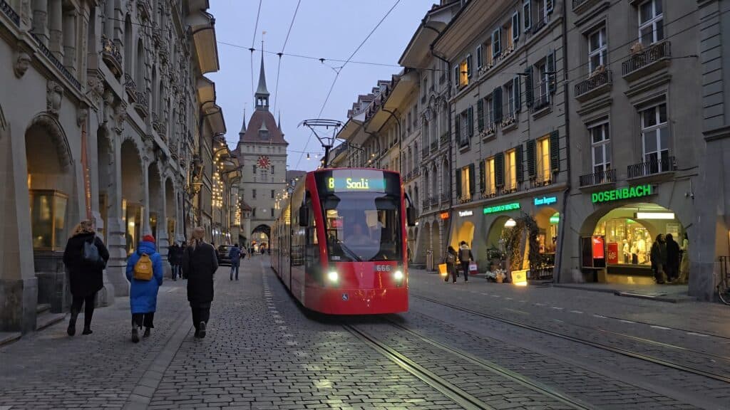 A vibrant street scene in Bern's historic UNESCO-listed Old Town, featuring the famous covered shopping arcades, local shops, and a modern tram passing through, as captured by author Ila from Tripilare.