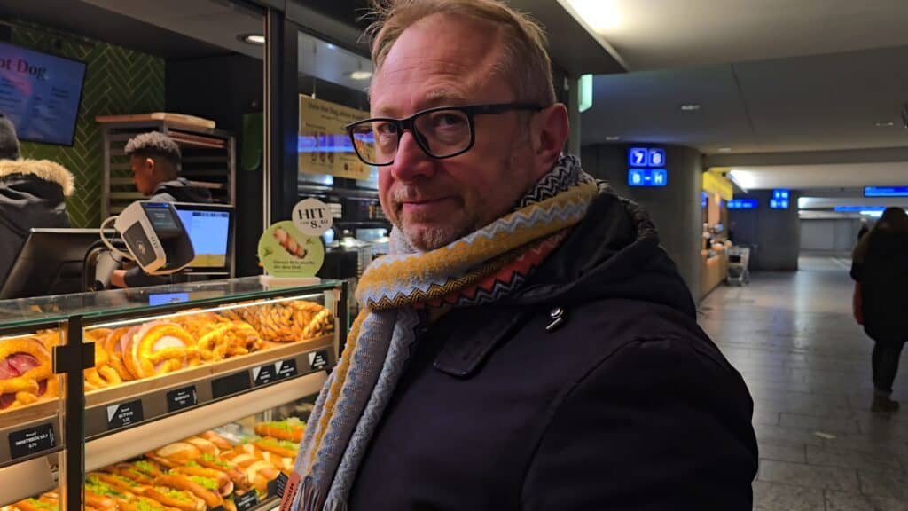 Gabriel, traveling with author Ila from Tripilare, standing in front of a Bretzelkönig kiosk at Bern station, enjoying a fresh pretzel before the return journey to Domodossola.