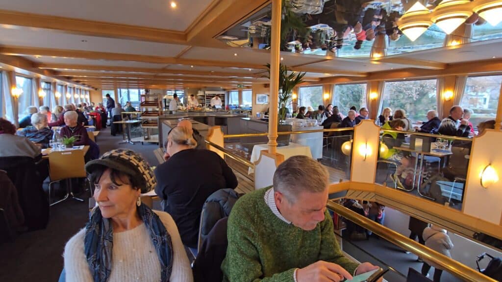 A wide shot of the crowded first-class interior of a Lake Thun boat, showing all tables occupied by passengers and at the lower floor people standing, including families with strollers, as documented by Ila from Tripilare.