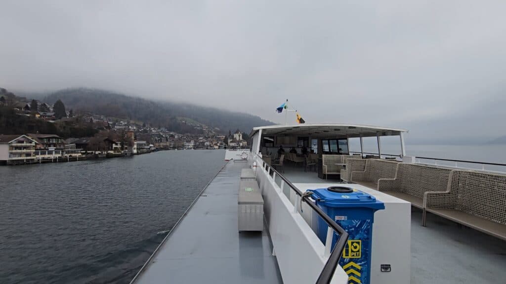 The outdoor deck of a Lake Thun ferry with empty seats due to the biting cold and thick fog obscuring the mountain scenery, as documented by Ila from Tripilare.