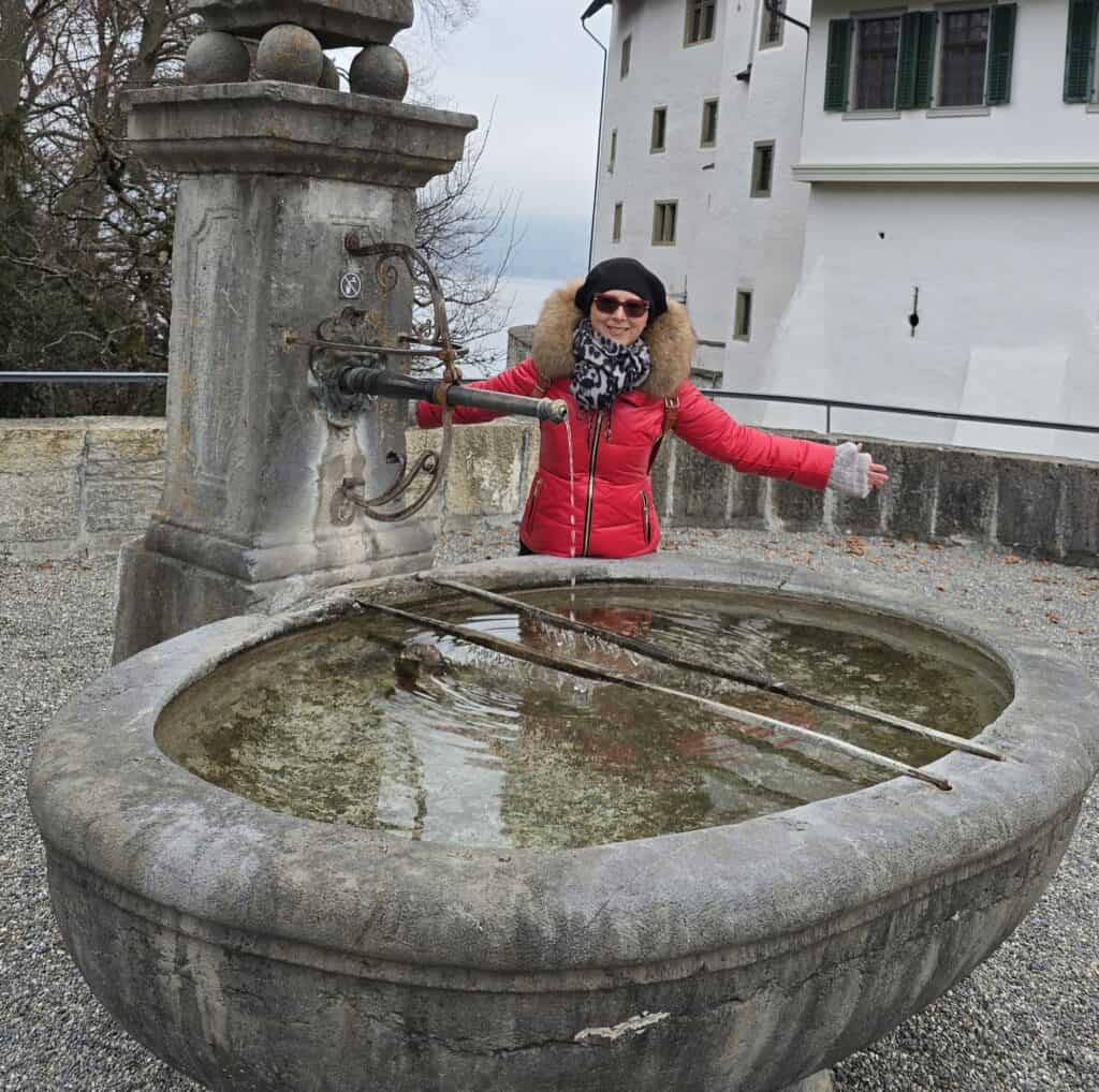 Author Ila from Tripilare standing by a stone fountain with partially frozen water outside the closed Spiez Castle, wearing heavy winter clothing in December 2025.