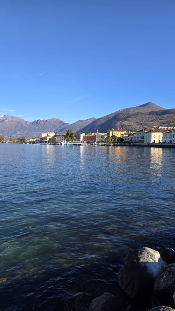 Panoramic vertical shot of Iseo, Italy, on a clear winter day. The colorful town buildings line the shore of the deep blue Lake Iseo, backed by mountains without snow. The foreground shows clear water over submerged rocks.