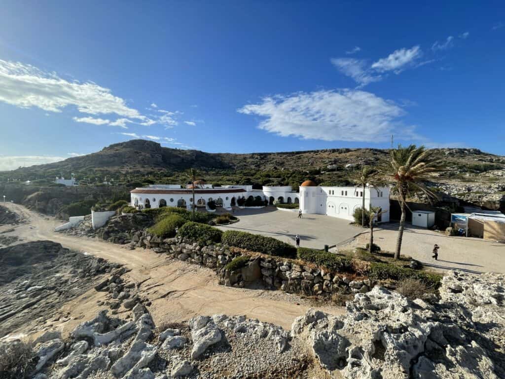 Majesty Reborn: Kallithea Springs Rhodes โ Italian Thermal Baths, Beaches & Architecture 4 Elevated panoramic view of the Kallithea Springs white architectural complex, showing the curved structure with multiple arches and red dome, set against the rocky, arid hills of Rhodes.