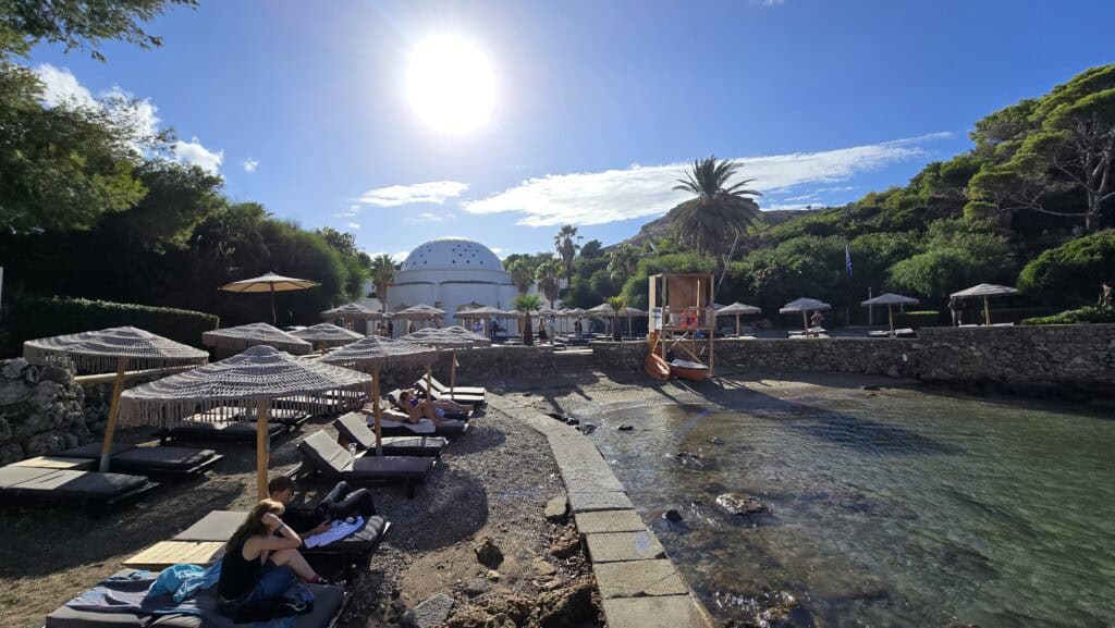 Majesty Reborn: Kallithea Springs Rhodes โ Italian Thermal Baths, Beaches & Architecture 8 Kallithea Springs beach area with dark sunbeds and straw umbrellas, showing the iconic white-domed Rotunda building and clear water on a sunny day in Rhodes.