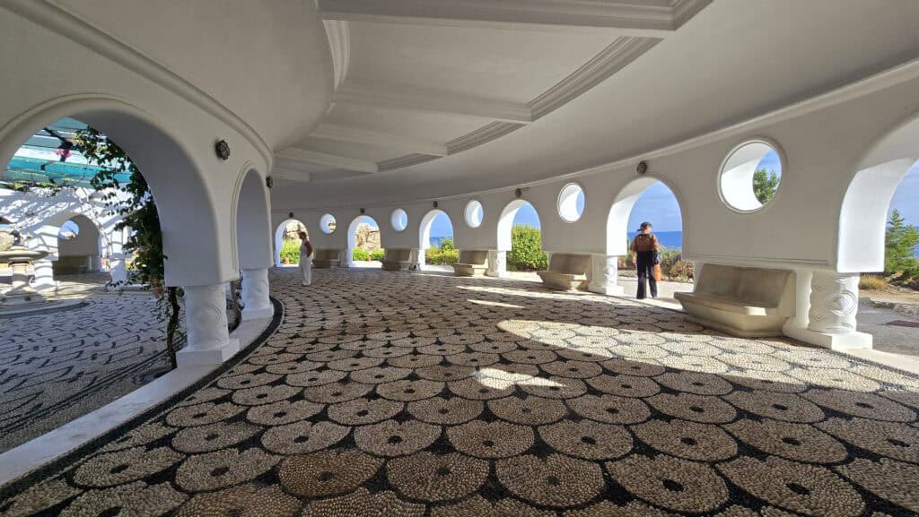 Majesty Reborn: Kallithea Springs Rhodes โ Italian Thermal Baths, Beaches & Architecture 1 Curved Art Deco portico at Kallithea Springs with white columns and arches, featuring intricate circular pebble mosaics and a panoramic view of the deep blue Aegean Sea.