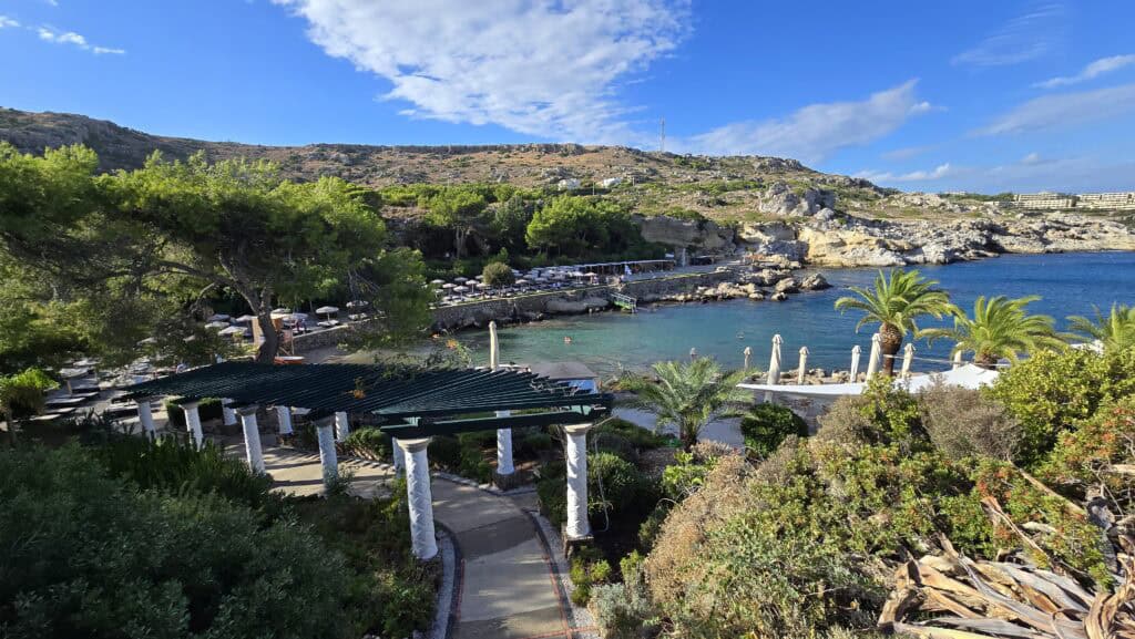Majesty Reborn: Kallithea Springs Rhodes โ Italian Thermal Baths, Beaches & Architecture 3 Panoramic view of Kallithea Springs pathway leading through lush Mediterranean gardens with white columns and a pergola, showcasing the cove, clear sea, and rocky coastline.
