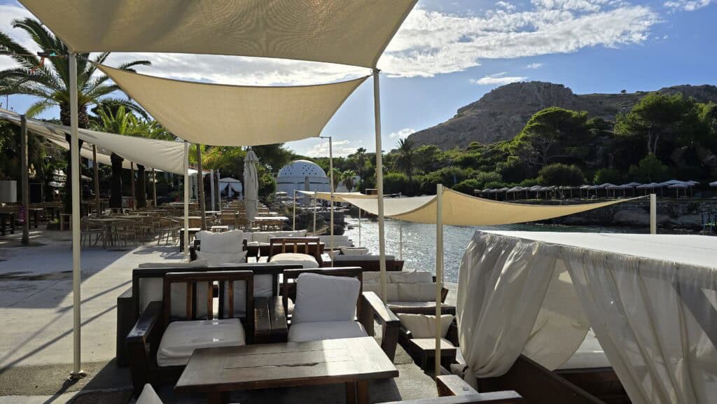 Majesty Reborn: Kallithea Springs Rhodes โ Italian Thermal Baths, Beaches & Architecture 9 Empty outdoor lounge seating area and tables at the Kallithea Springs beach bar, featuring white cushions and sun shades, overlooking the sea and a distant hill.